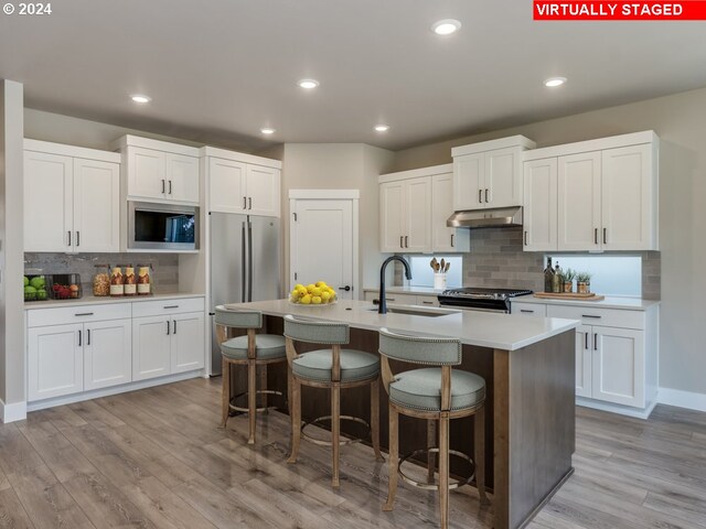 kitchen with stainless steel appliances, decorative backsplash, a center island with sink, white cabinets, and light wood-type flooring