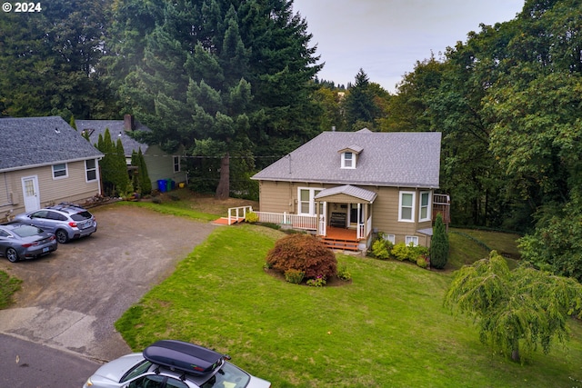 view of front facade featuring roof with shingles, a front yard, a deck, a view of trees, and driveway