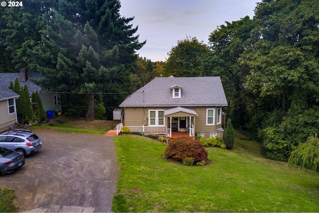 view of front facade featuring roof with shingles, covered porch, a view of trees, driveway, and a front lawn