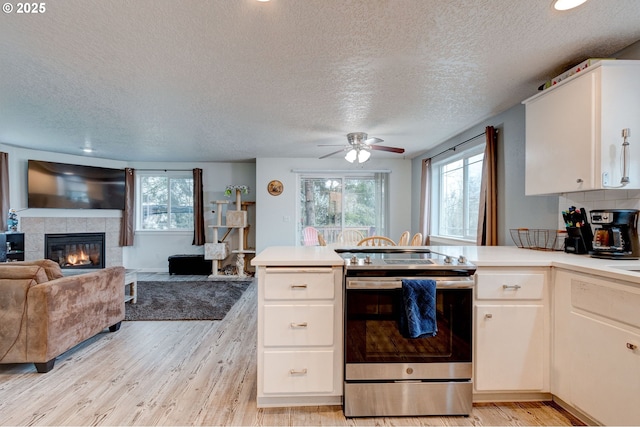 kitchen featuring a tile fireplace, stainless steel electric stove, white cabinets, ceiling fan, and a textured ceiling