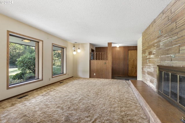 unfurnished living room with a textured ceiling, a stone fireplace, visible vents, carpet floors, and wooden walls
