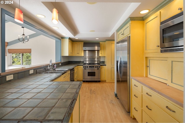 kitchen with sink, hanging light fixtures, stainless steel appliances, tile counters, and wall chimney exhaust hood