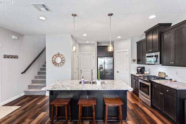 kitchen featuring sink, decorative light fixtures, appliances with stainless steel finishes, an island with sink, and light stone countertops