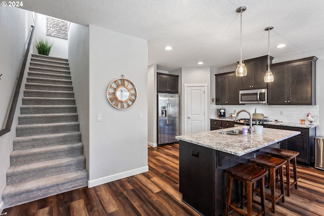 kitchen with dark wood-type flooring, appliances with stainless steel finishes, light stone countertops, an island with sink, and a textured ceiling