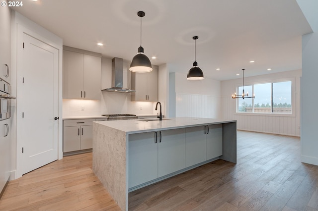 kitchen with wall chimney exhaust hood, an island with sink, sink, decorative light fixtures, and light hardwood / wood-style floors