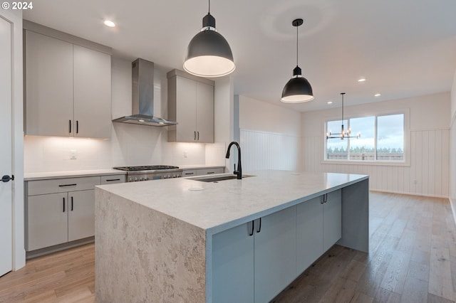 kitchen featuring sink, light wood-type flooring, wall chimney exhaust hood, decorative light fixtures, and a center island with sink