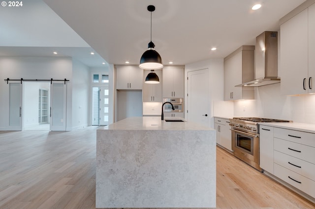 kitchen featuring hanging light fixtures, a barn door, light hardwood / wood-style flooring, wall chimney exhaust hood, and stainless steel appliances