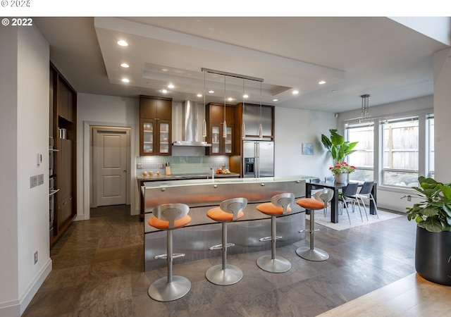 kitchen with wall chimney exhaust hood, stainless steel fridge, pendant lighting, a tray ceiling, and a kitchen bar