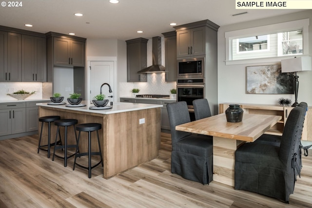 kitchen with a kitchen breakfast bar, light wood-type flooring, wall chimney exhaust hood, stainless steel appliances, and a center island with sink