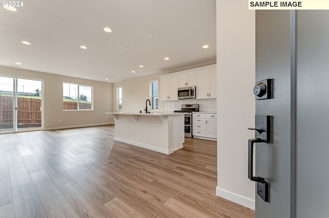 kitchen with light hardwood / wood-style flooring, appliances with stainless steel finishes, white cabinetry, backsplash, and an island with sink