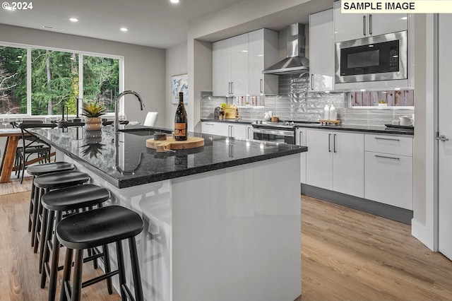 kitchen featuring stainless steel stove, light hardwood / wood-style floors, an island with sink, wall chimney exhaust hood, and built in microwave