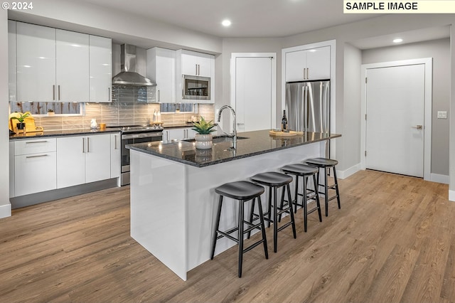 kitchen with wall chimney range hood, light hardwood / wood-style floors, a kitchen island with sink, white cabinetry, and appliances with stainless steel finishes