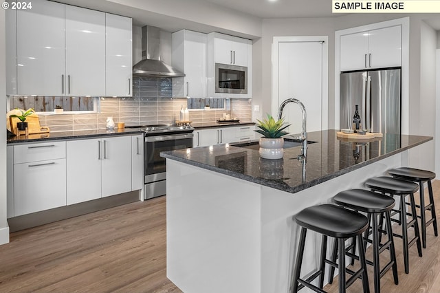 kitchen featuring wall chimney range hood, stainless steel appliances, white cabinetry, an island with sink, and light wood-type flooring