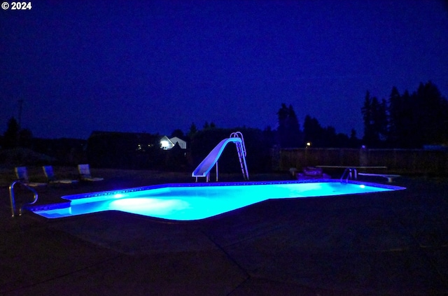 pool at night featuring a diving board, a water slide, and a fenced in pool