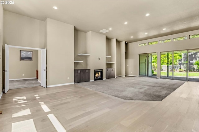 unfurnished living room with a high ceiling, recessed lighting, baseboards, wood finished floors, and a lit fireplace