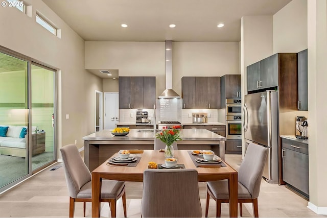 kitchen featuring stainless steel appliances, backsplash, light wood finished floors, wall chimney range hood, and a kitchen island with sink