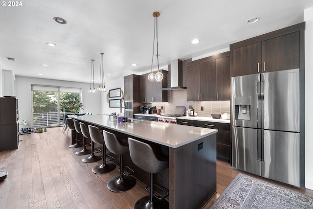 kitchen featuring wall chimney range hood, dark hardwood / wood-style floors, stainless steel fridge, decorative light fixtures, and a center island with sink