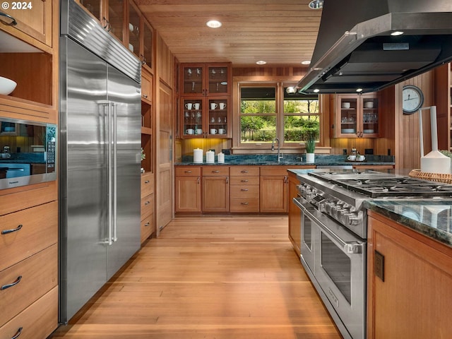 kitchen with high end appliances, wood ceiling, glass insert cabinets, dark stone countertops, and island exhaust hood