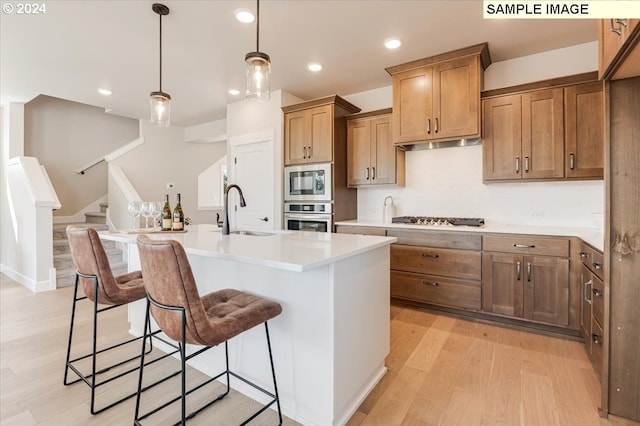 kitchen with a center island with sink, hanging light fixtures, sink, light wood-type flooring, and appliances with stainless steel finishes