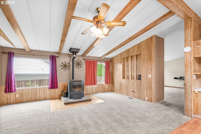 unfurnished living room featuring wooden walls, a wood stove, carpet floors, ceiling fan, and vaulted ceiling with beams