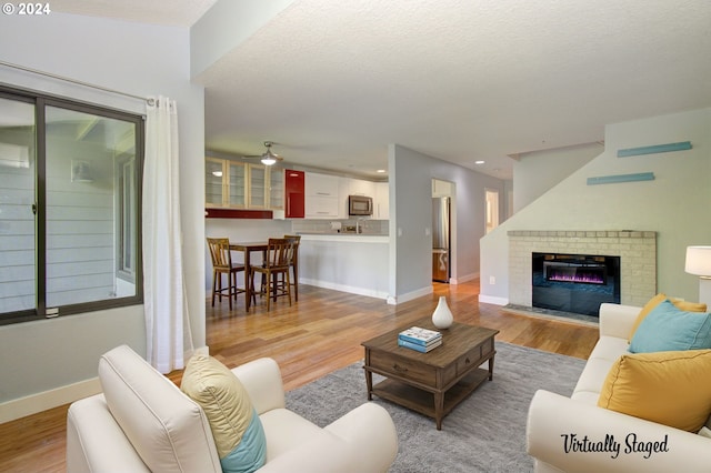 living room with a brick fireplace, a textured ceiling, light hardwood / wood-style flooring, and ceiling fan