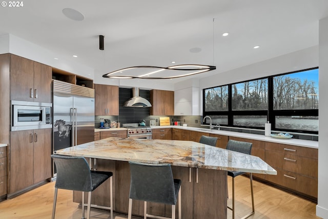 kitchen featuring sink, wall chimney range hood, backsplash, built in appliances, and light wood-type flooring