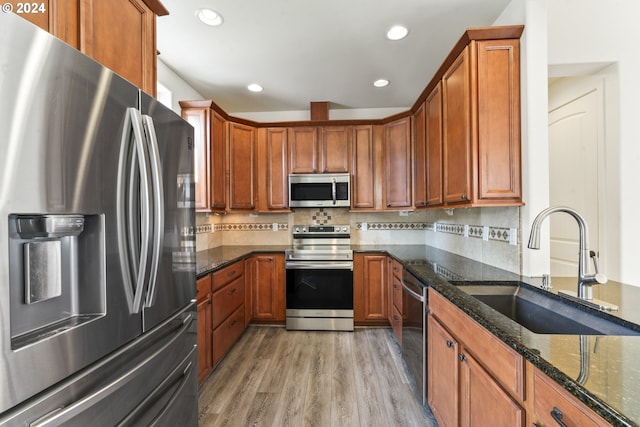 kitchen with a sink, appliances with stainless steel finishes, brown cabinets, dark stone counters, and light wood finished floors