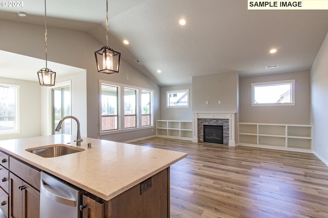 kitchen with a stone fireplace, sink, hanging light fixtures, light wood-type flooring, and an island with sink