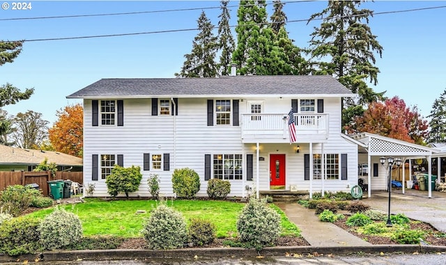 colonial-style house featuring a carport, a front lawn, a balcony, and fence