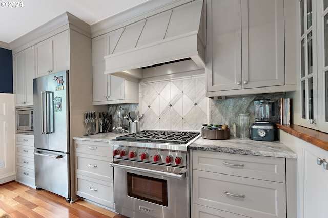 kitchen with high quality appliances, backsplash, light stone counters, custom range hood, and light wood-type flooring