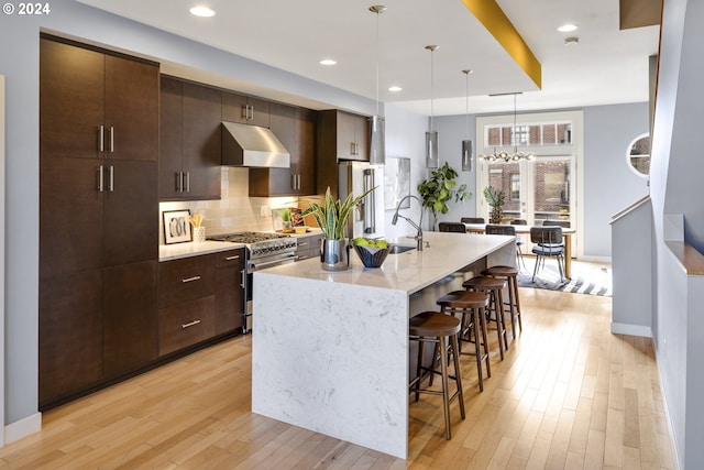 kitchen featuring decorative light fixtures, an island with sink, dark brown cabinets, range hood, and appliances with stainless steel finishes