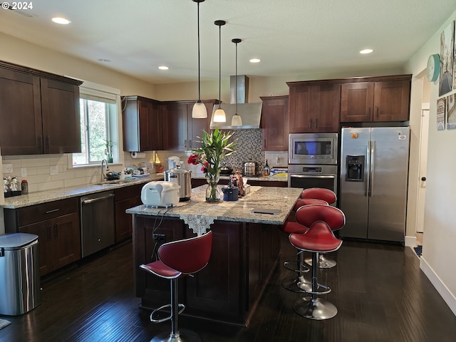 kitchen with light stone counters, ventilation hood, a kitchen island, pendant lighting, and appliances with stainless steel finishes