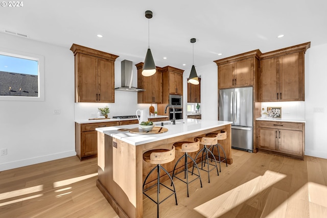 kitchen with a kitchen bar, light wood-type flooring, a kitchen island with sink, wall chimney range hood, and built in appliances