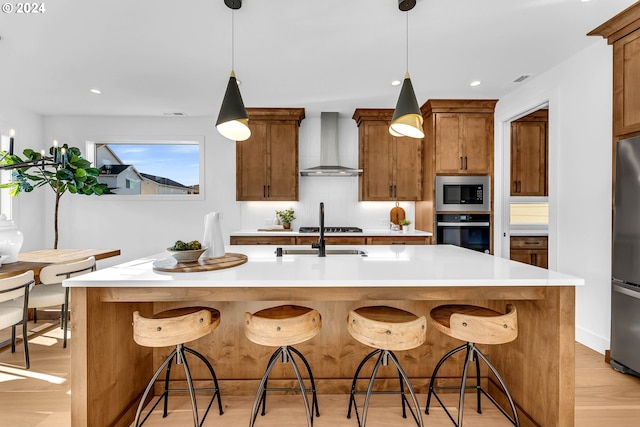 kitchen featuring a kitchen bar, appliances with stainless steel finishes, wall chimney range hood, light hardwood / wood-style floors, and an island with sink