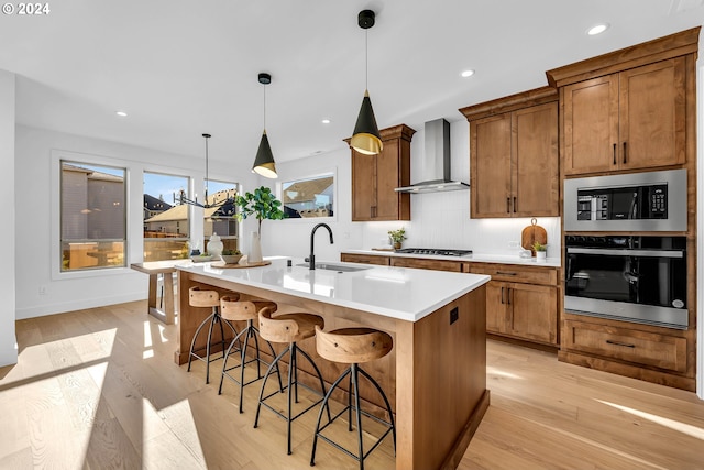 kitchen with stainless steel appliances, a kitchen island with sink, wall chimney range hood, decorative light fixtures, and light hardwood / wood-style floors