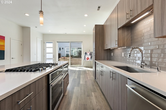 kitchen with backsplash, stainless steel appliances, sink, hanging light fixtures, and light hardwood / wood-style floors