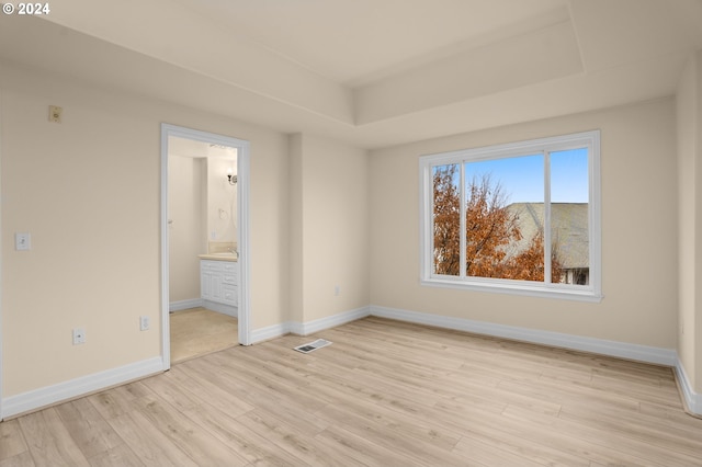 unfurnished bedroom with visible vents, baseboards, a raised ceiling, ensuite bath, and light wood-type flooring