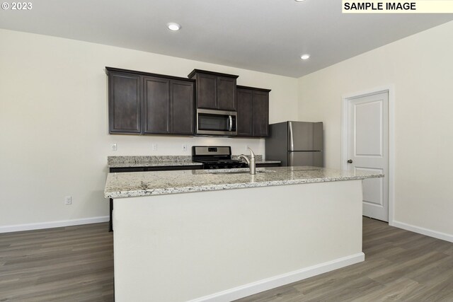 kitchen featuring an island with sink, wood finished floors, stainless steel appliances, dark brown cabinetry, and baseboards