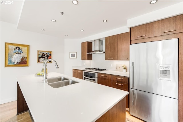 kitchen with wall chimney exhaust hood, stainless steel appliances, sink, light hardwood / wood-style floors, and an island with sink