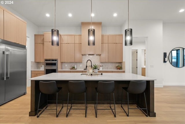 kitchen featuring light brown cabinetry, appliances with stainless steel finishes, a center island with sink, and hanging light fixtures