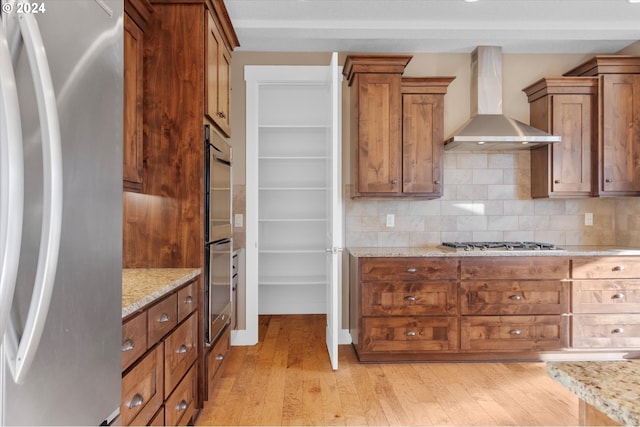 kitchen with backsplash, light stone counters, stainless steel appliances, wall chimney range hood, and light hardwood / wood-style floors