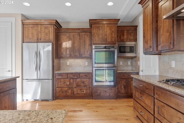 kitchen featuring wall chimney exhaust hood, light stone counters, light hardwood / wood-style flooring, decorative backsplash, and appliances with stainless steel finishes