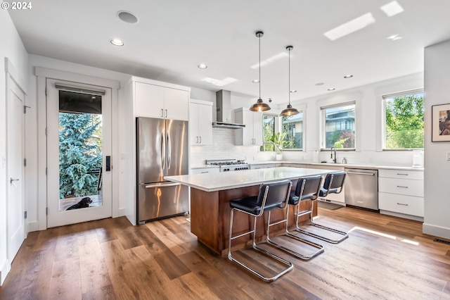 kitchen featuring appliances with stainless steel finishes, white cabinets, wall chimney range hood, decorative light fixtures, and a kitchen island