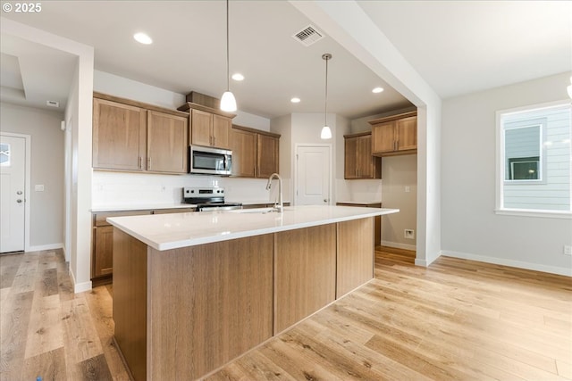 kitchen with appliances with stainless steel finishes, a kitchen island with sink, hanging light fixtures, and light hardwood / wood-style flooring