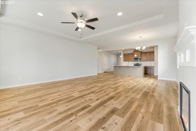 unfurnished living room featuring a tray ceiling, a tiled fireplace, ceiling fan with notable chandelier, and light wood-type flooring