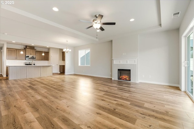 unfurnished living room with a tray ceiling, ceiling fan with notable chandelier, a tile fireplace, and light wood-type flooring