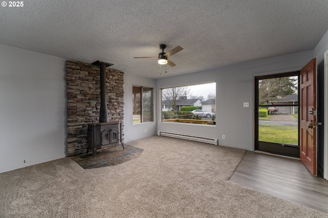 unfurnished living room with a textured ceiling, ceiling fan, a baseboard heating unit, carpet floors, and a wood stove