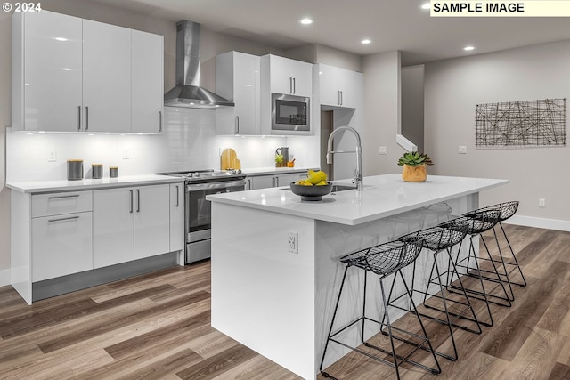 kitchen with white cabinetry, appliances with stainless steel finishes, wall chimney range hood, and an island with sink