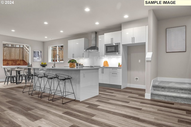 kitchen with a center island with sink, a kitchen bar, wall chimney exhaust hood, stainless steel microwave, and white cabinets