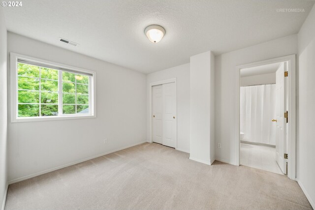 unfurnished bedroom with light colored carpet, a textured ceiling, a closet, and ensuite bathroom